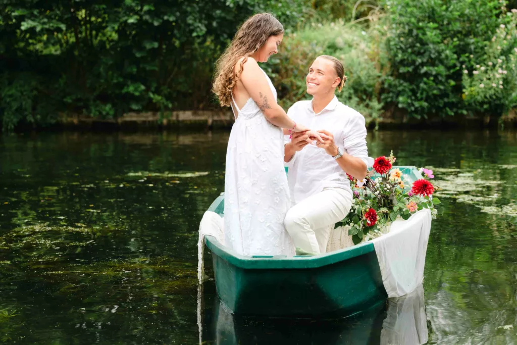 jeune femme souriante, jeune homme chemise blanche, demande, bague de fiançailles, coupe de champagne, barque fleurie, hortillonnages Amiens, séance photo couple, romance au fil de l’eau, demande en mariage, photographe Amiens, expérience romantique, moment complice, fleurs colorées, amour authentique, Hauts-de-France, souvenir romantique, photographie naturelle, barque traditionnelle, expérience poétique
