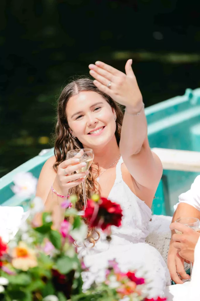 jeune femme souriante, robe blanche, bague de fiançailles, coupe de champagne, barque fleurie, hortillonnages Amiens, séance photo couple, romance au fil de l’eau, demande en mariage, photographe Amiens, expérience romantique, moment complice, fleurs colorées, amour authentique, Hauts-de-France, souvenir romantique, photographie naturelle, barque traditionnelle, expérience poétique